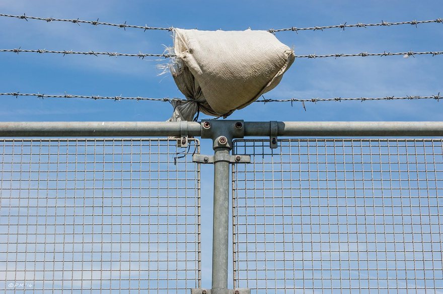 Sand bag draped over barbed wire on top of wire mesh fence against blue sky & light cloud, abstract urban, Brighton 2014 eyeteeth.net