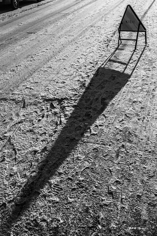 Warning sign stands on snow covered road casting long shadow. Landport, Lewes, East Sussex UK. Monochrome Landscape. © P. Maton 2015 eyeteeth.net