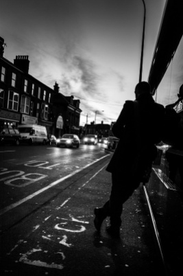 Silhouette of man leaning against bus stop shelter next to road with cars illuminating street and dawn light over Preston Circus, Brighton UK. Monochrome black and white urban street photography. © P. Maton 2016 eyeteeth.net