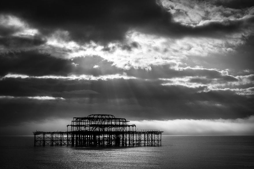 Ruined framework of abandoned West Pier Brighton UK with dramatic clouds and sun rays shining on water. Black and white seascape, moody atmospheric. © P, Maton 2014 eyeteeth.net