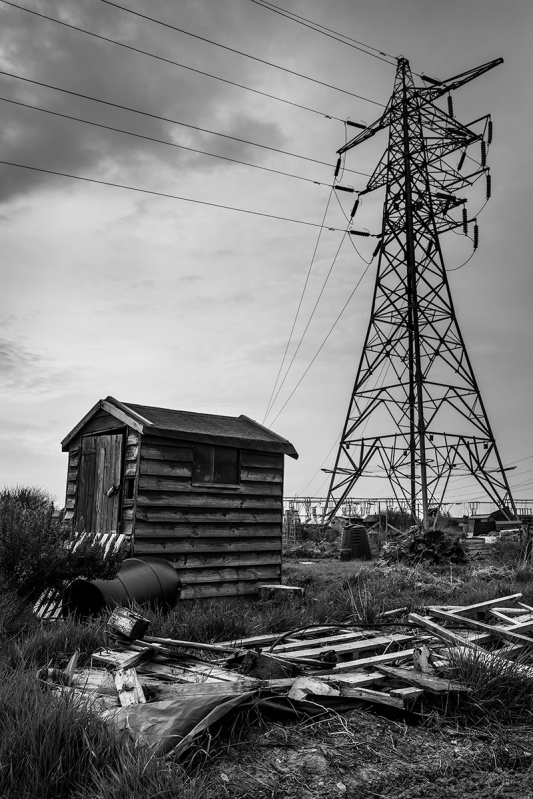 Allotment shed with electricity pylon in background and smashed wooden pallet in foreground. Black and white documentary photograph portrait urban Britain. Fujifilm XT-2 XF 16mm 1.4 R WR. Portslade UK. © P. Maton 2017 eyeteeth.net