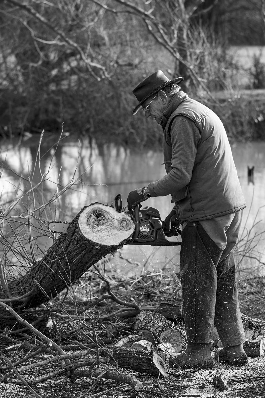 man in hat sawing Willow with a chainsaw, side profile black and white rural environmental portrait Gabriel's Fisheries Kent UK ©P. Maton 2019 eyeteeth.net