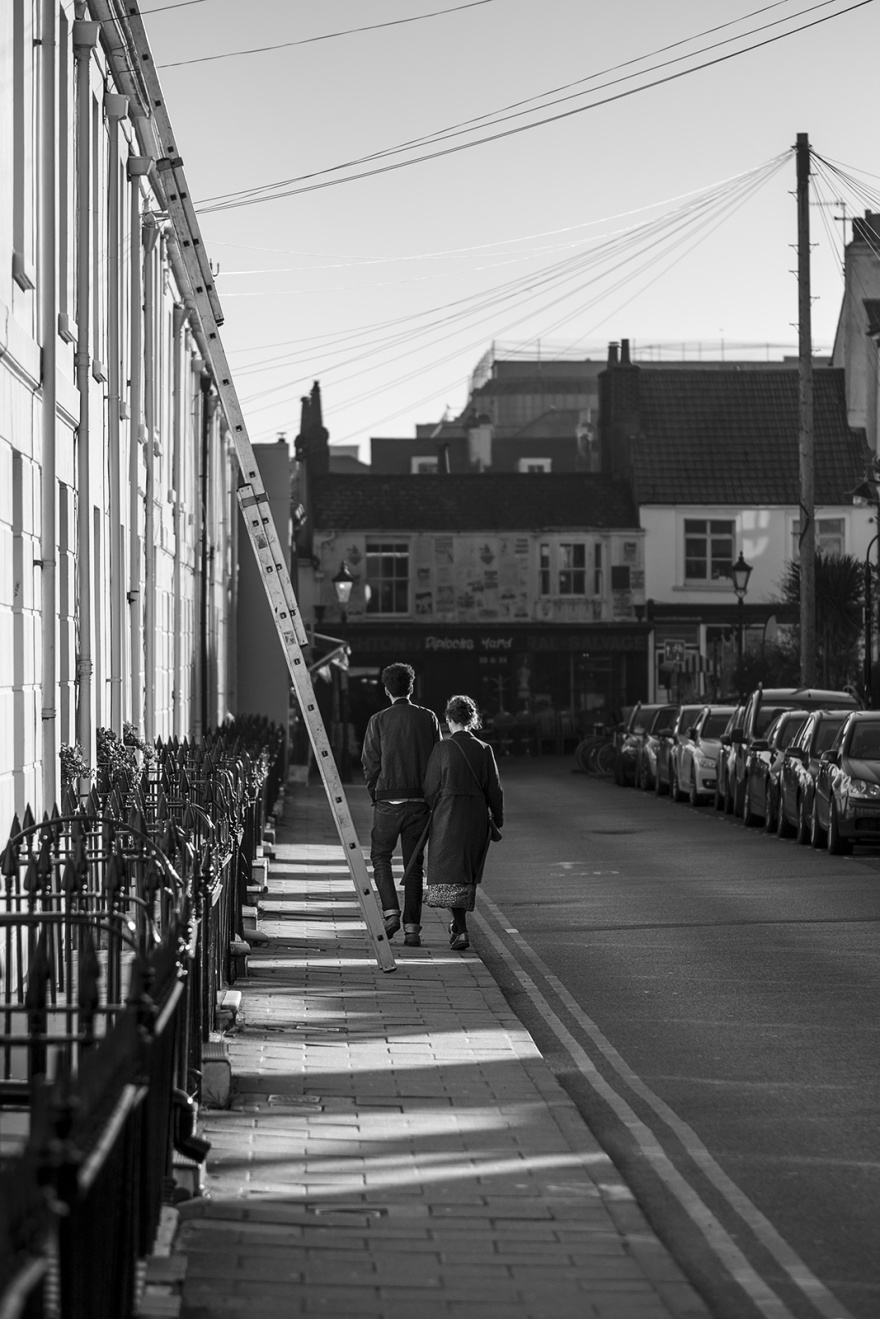 Young couple with linked arms walking away beside ladder down victorian town street, Kensington Place Brighton UK black and white documentary street photography portrait ©P. Maton 2019 eyeteeth.net